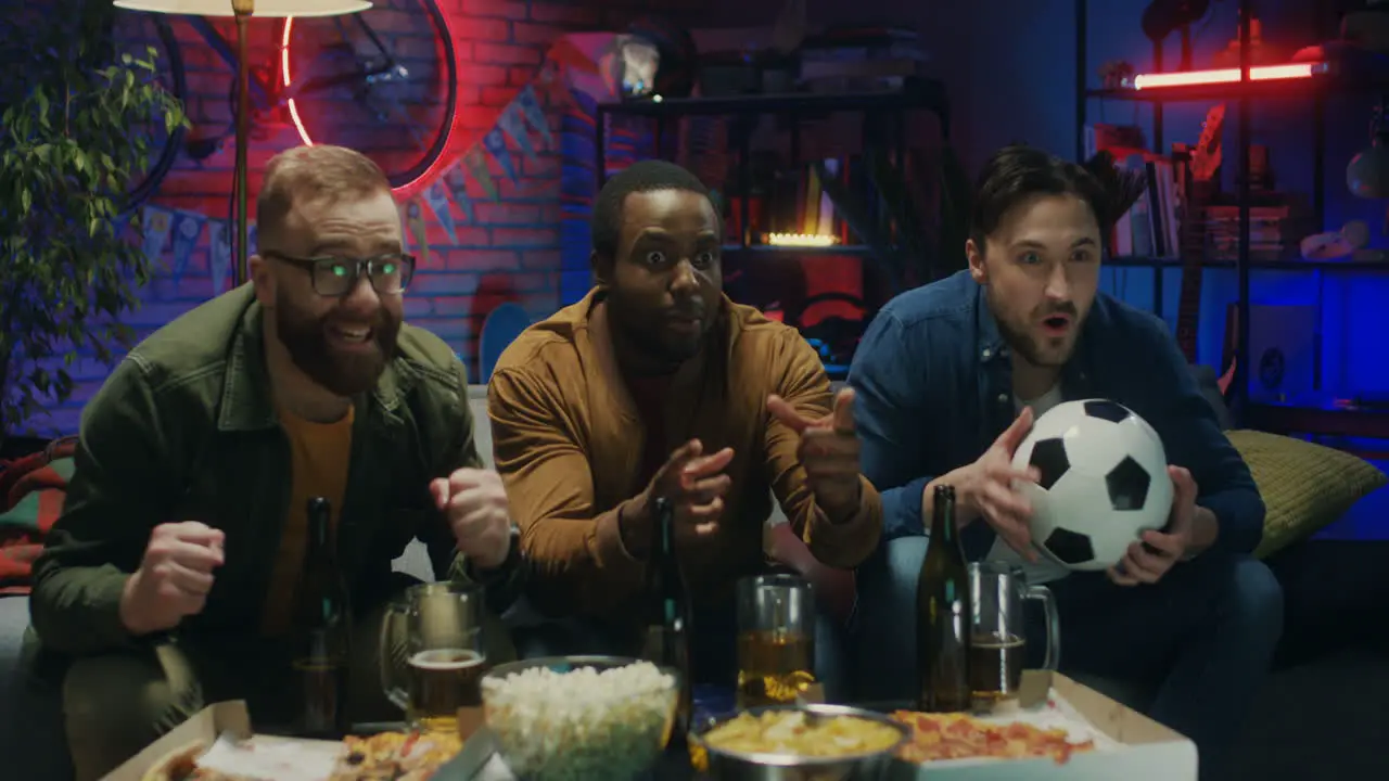 Three Multiethnic Friendly And Cheerful Young Men Celebrating Victory Of Their Football Team In The Championship In The Dark Living Room With Snacks And Beer