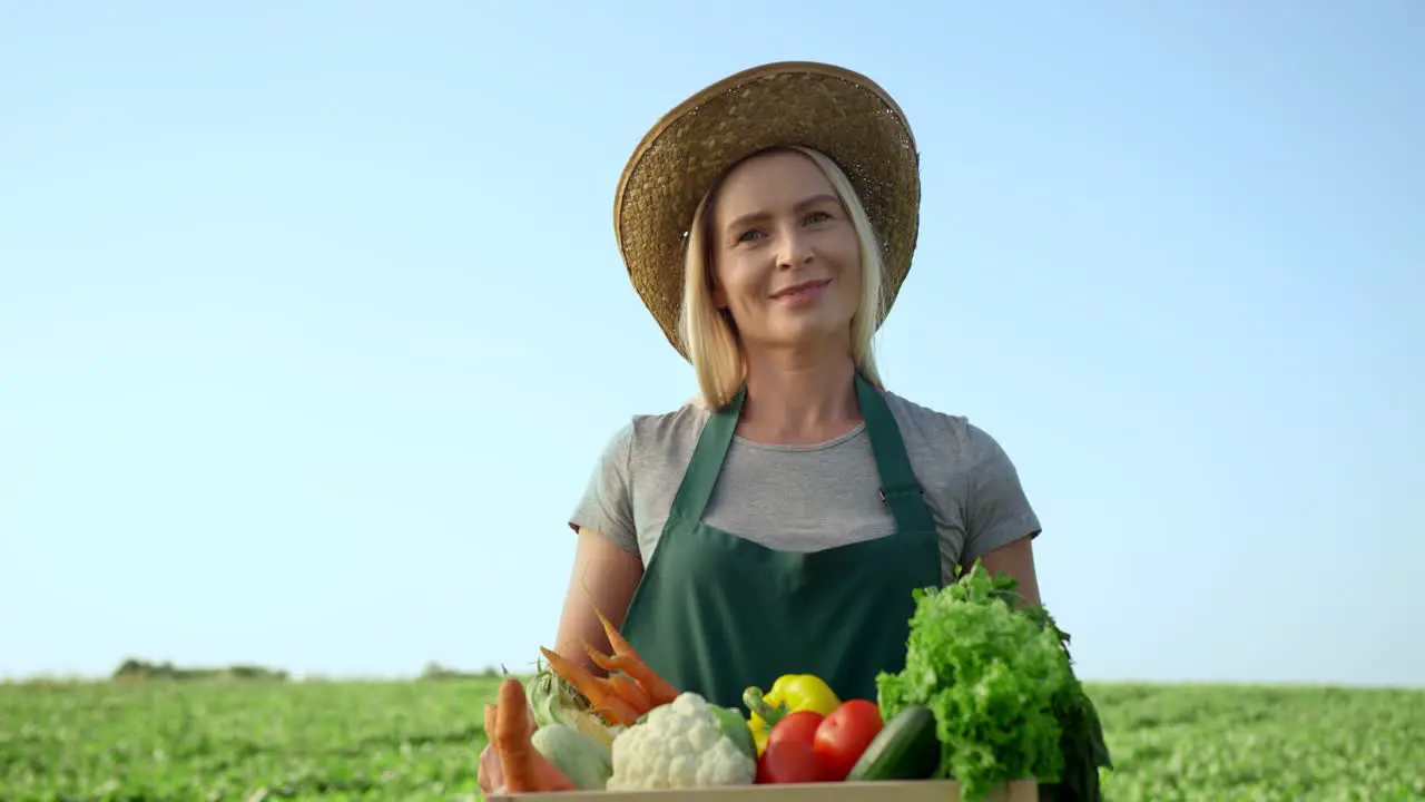 Portrait Shot Of The Blond Young Good Looking Woman Farmer In A Hat And With A Box Full Of The Harvested Vegetables In Field