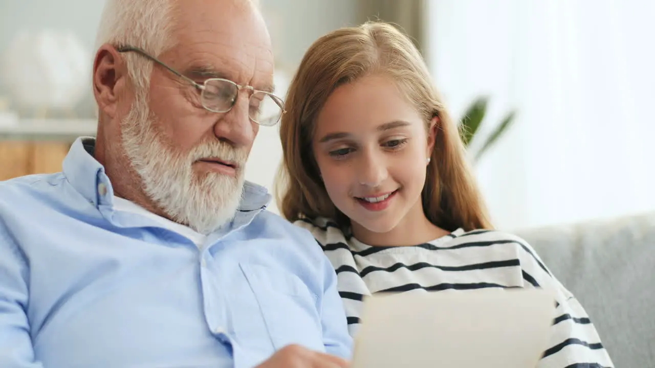 Close Up Of The Senior Grey Haired Grandpa In Glasses Demonstrating Old Photos To His Pretty Granddaughter While They Sitting On The Couch At Home