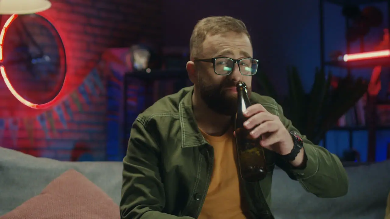 Portrait Shot Of The Happy Young Man In Glasses Watching Sport Channel On Tv In The Evening Drinking Beer And Smiling