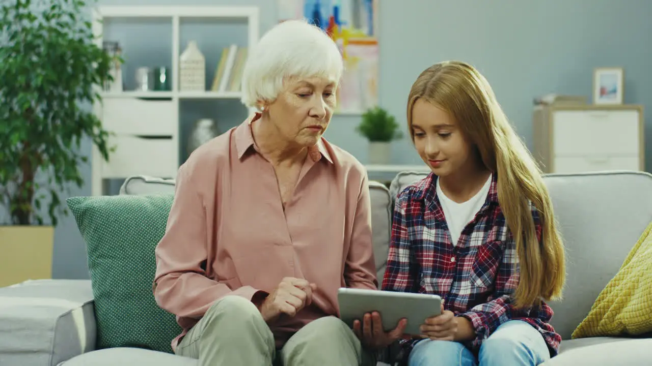 Pretty Teenager Girl Sitting On The Couch In The Nice Room Using Tablet Computer And Talking With Her Grandmother Who Sitting Next To Her