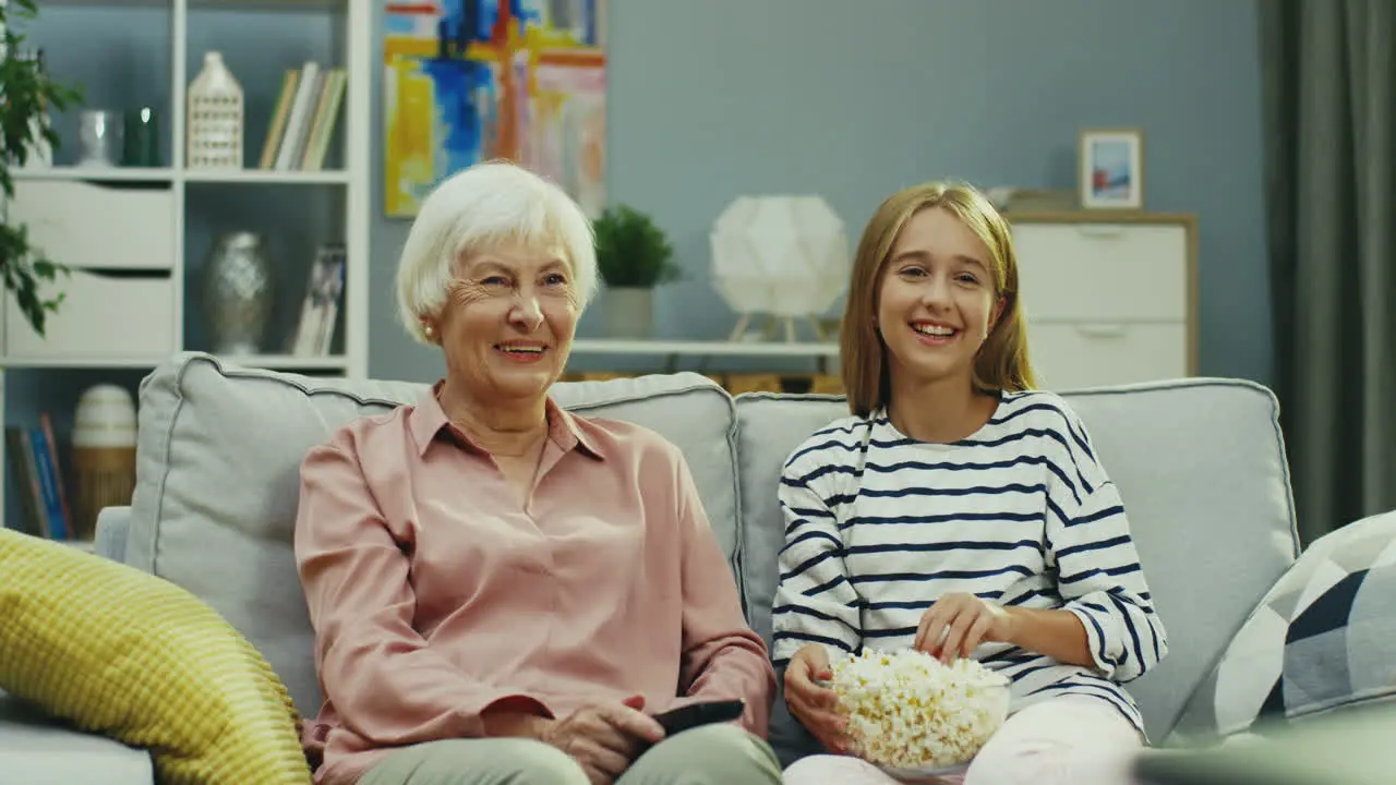 Portrait Shot Of The Pretty Small Teenager Girl And Senior Grandmother Sitting Together On The Sofa At Home Watching Tv And Laughing