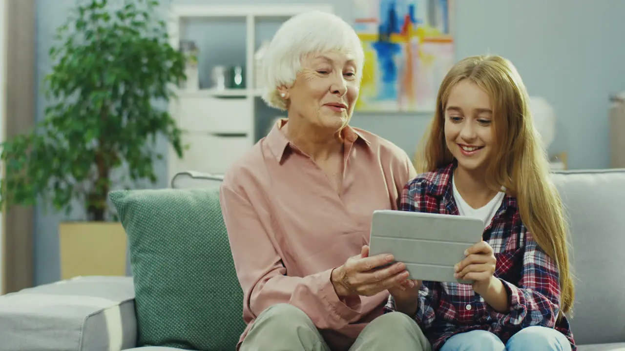 Cheerful Andsmiled Grandmother And Granddaughter Sitting Together On The Sofa At Home And Watching Something On The Tablet Device