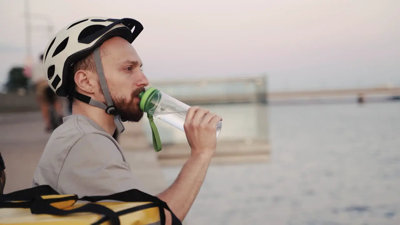 Food Delivery Guy Seated Next To A River At Sunset Drinks Some Water During His Break