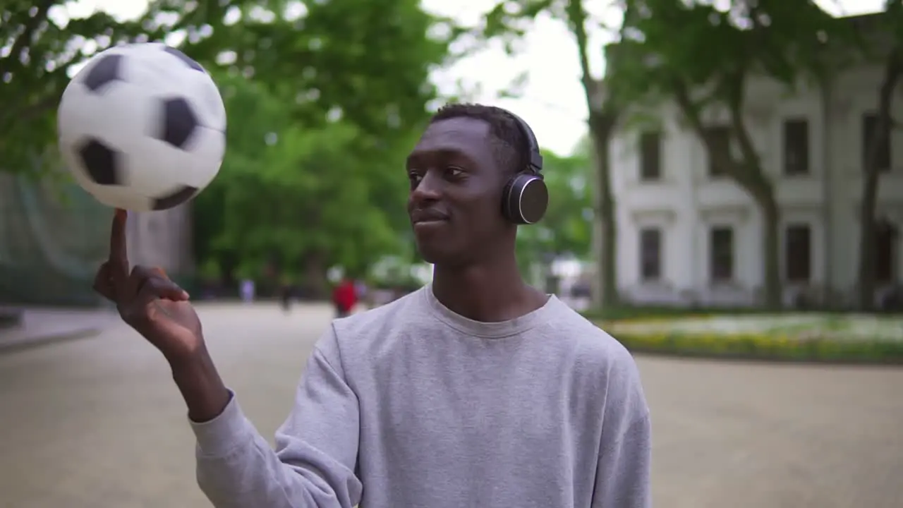 A Stylish Man In Casual Clothes And Black Headphones Is Spinning A Football Ball Above Him While Standing On City Street Alone