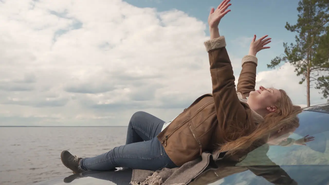 Red Haired Woman Lying On The Bonnet Of Her Car Next To A Beautiful Lake She Covers The Clouds With Her Hands