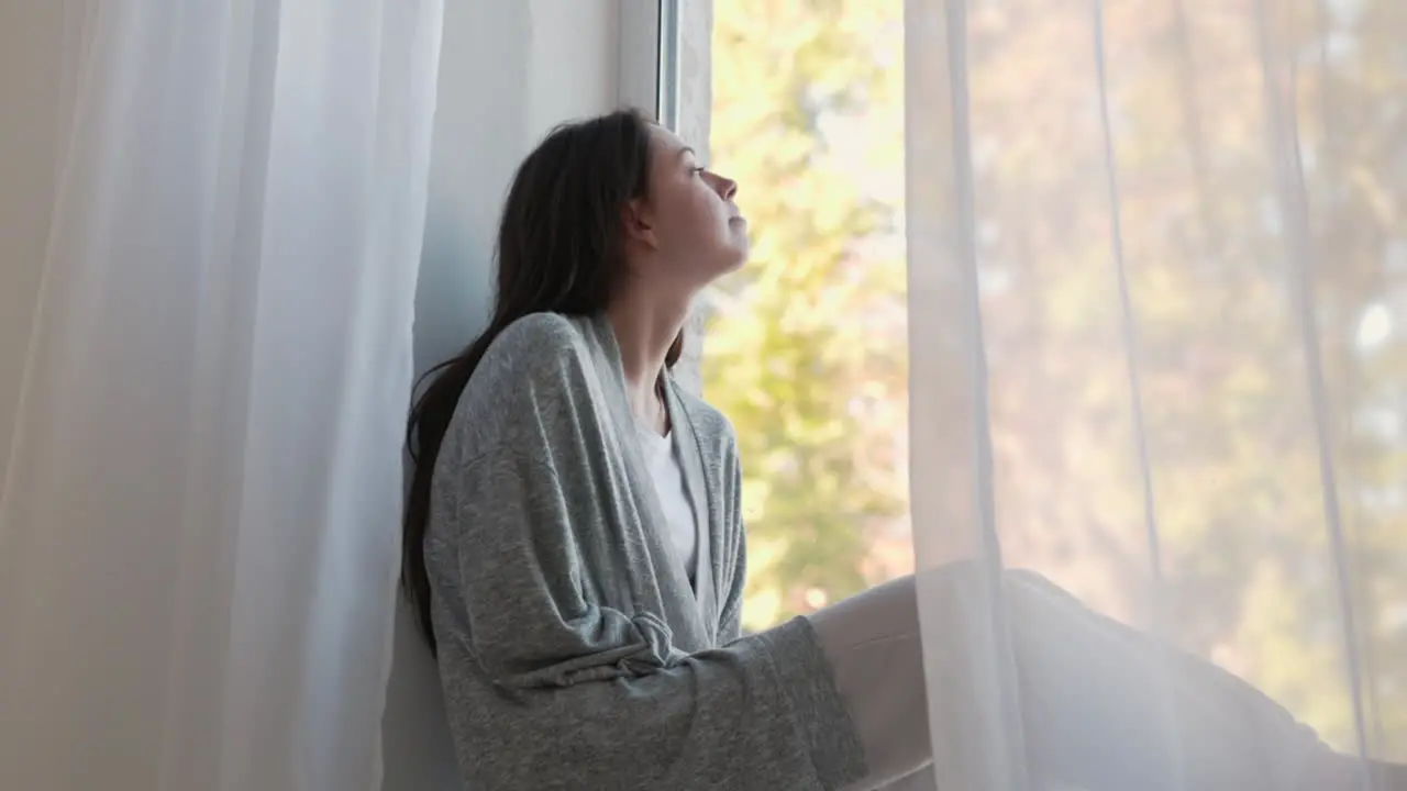 Pensive Woman Sitting On A Windowsill Looking Outside Through The Window In The Morning At Home