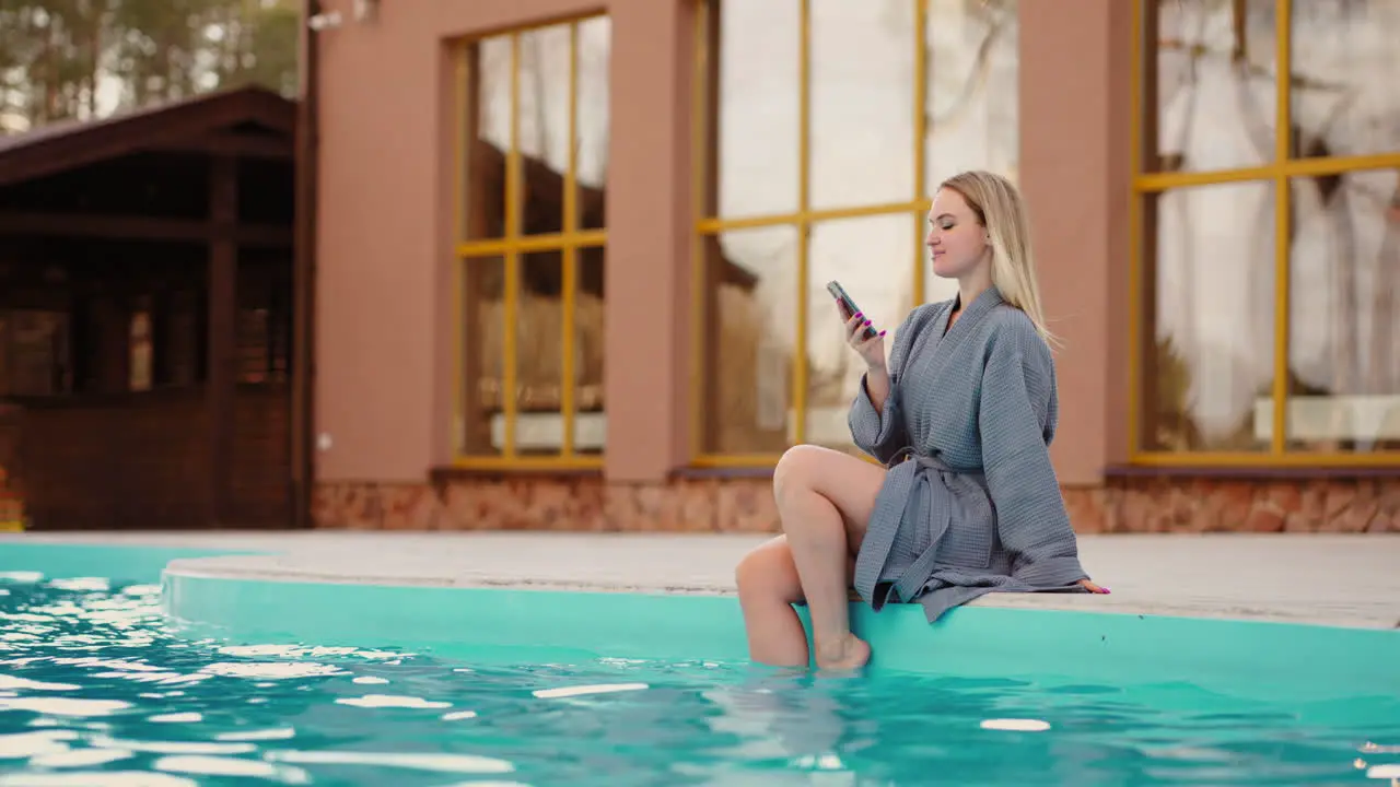 pretty woman is sittign on edge of outdoor thermal pool and resting surfing internet by modern smartphone