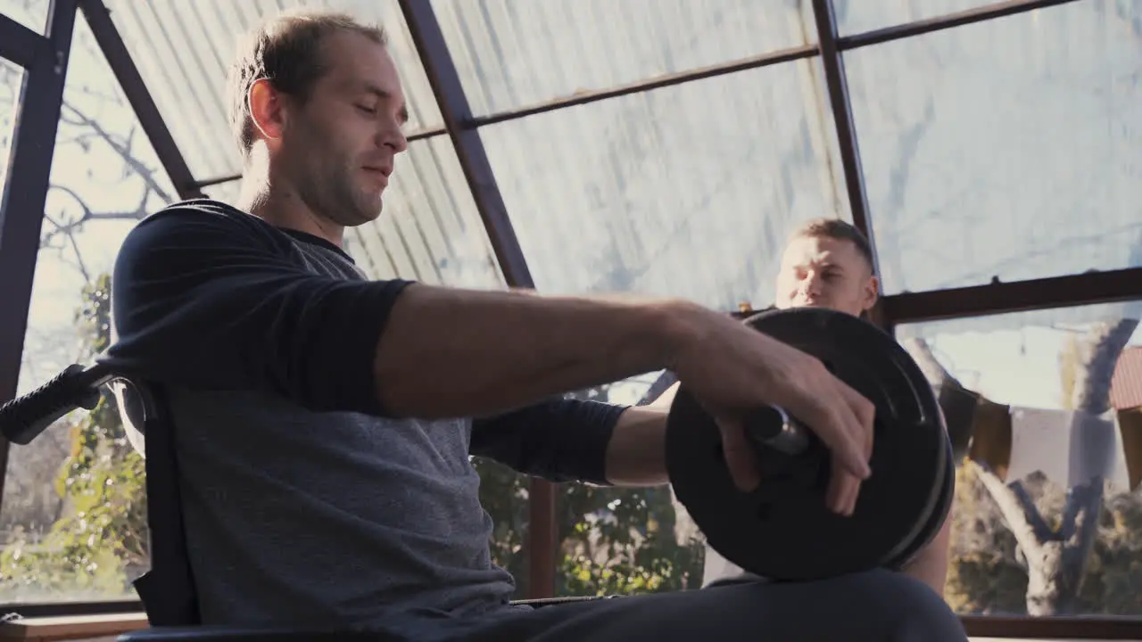 Side View Of A Disabled Man In Wheelchair And His Personal Trainer Working Out With Dumbbell At Home