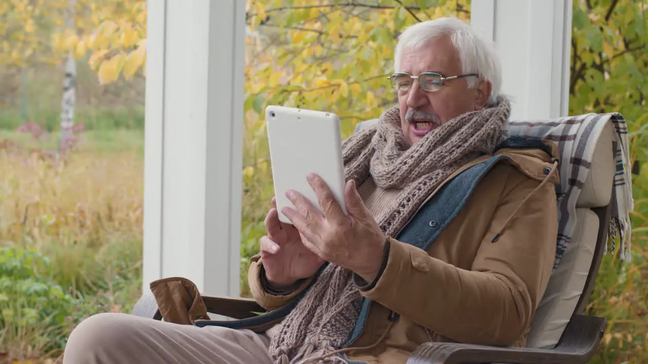 Senior Man Sitting On A Bench In Home Garden While Talking On A Video Call Using A Tablet