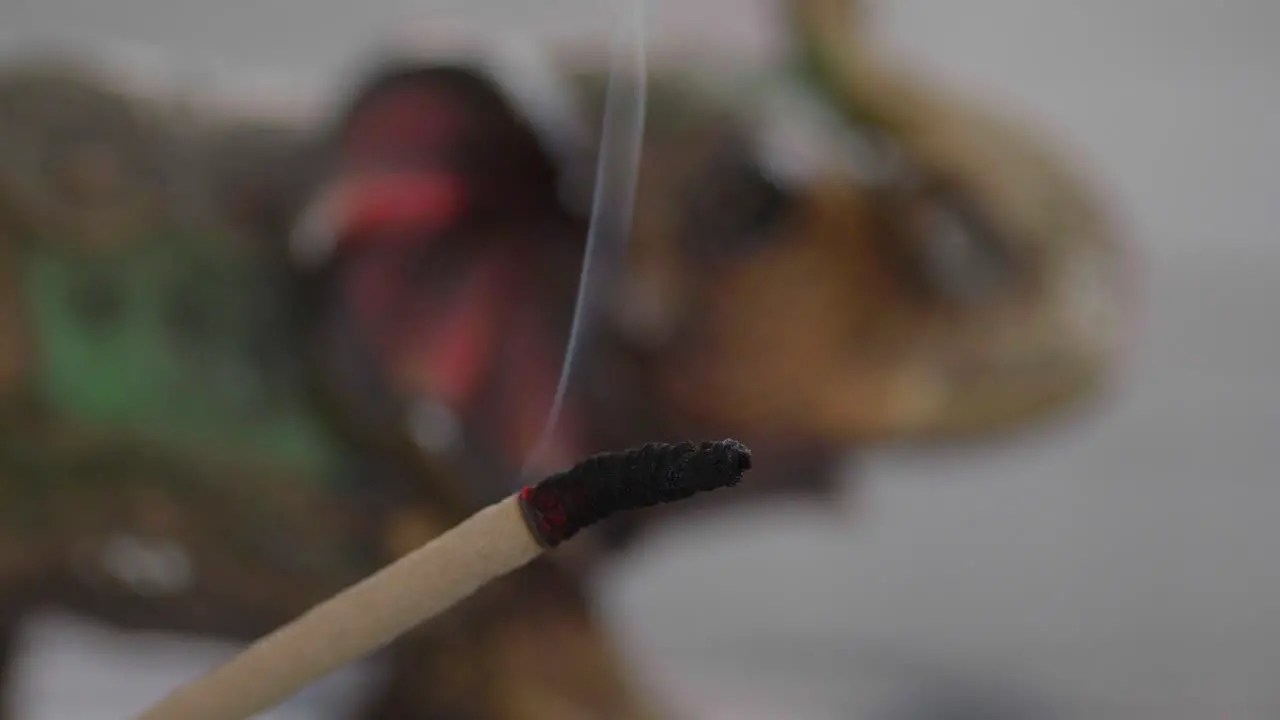 Close-up of a burning incense stick with an ornated elephant figure in the background