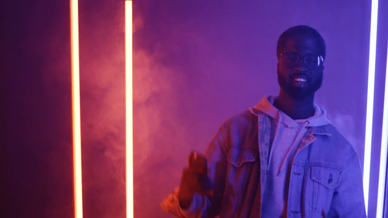 American Stylish Man In Glasses Dancing Holding Smartphone And Looking At Camera On Pink And Violet Neon Light Background