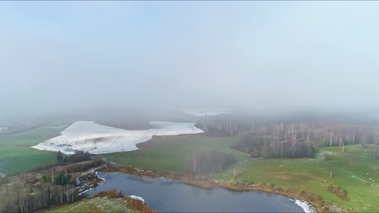 Aerial drone shot through white clouds over snow melting on a meadow alongside small lakes on an early spring day