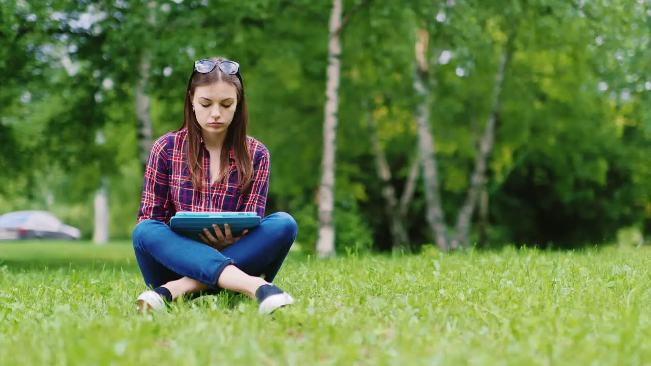 A Female Student Is Preparing For Exams Sits On A Lawn In The Park Enjoys A Tablet Hd Video