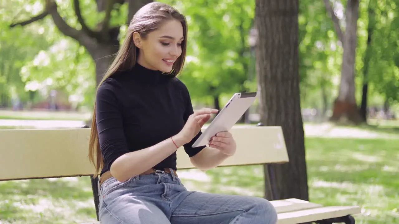 Smiling beautiful woman sitting on bench in green park and using tablet