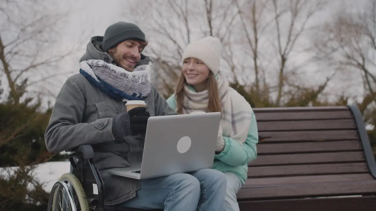 Disabled Man In Wheelchair And His Friend Watching Something Funny On Laptop Computer While Drinking Takeaway Coffee At Urban Park In Winter 4