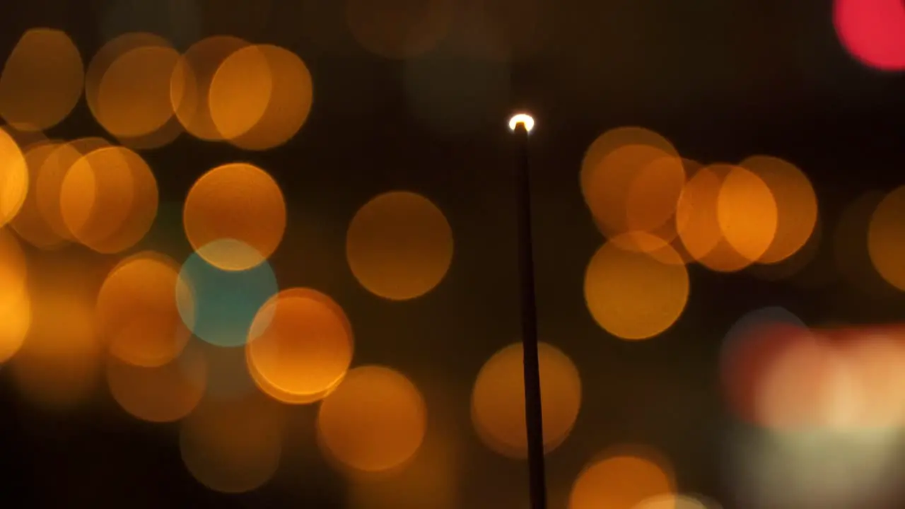 Still shot of a hand burning an incense stick with a blurry background with lights during the night