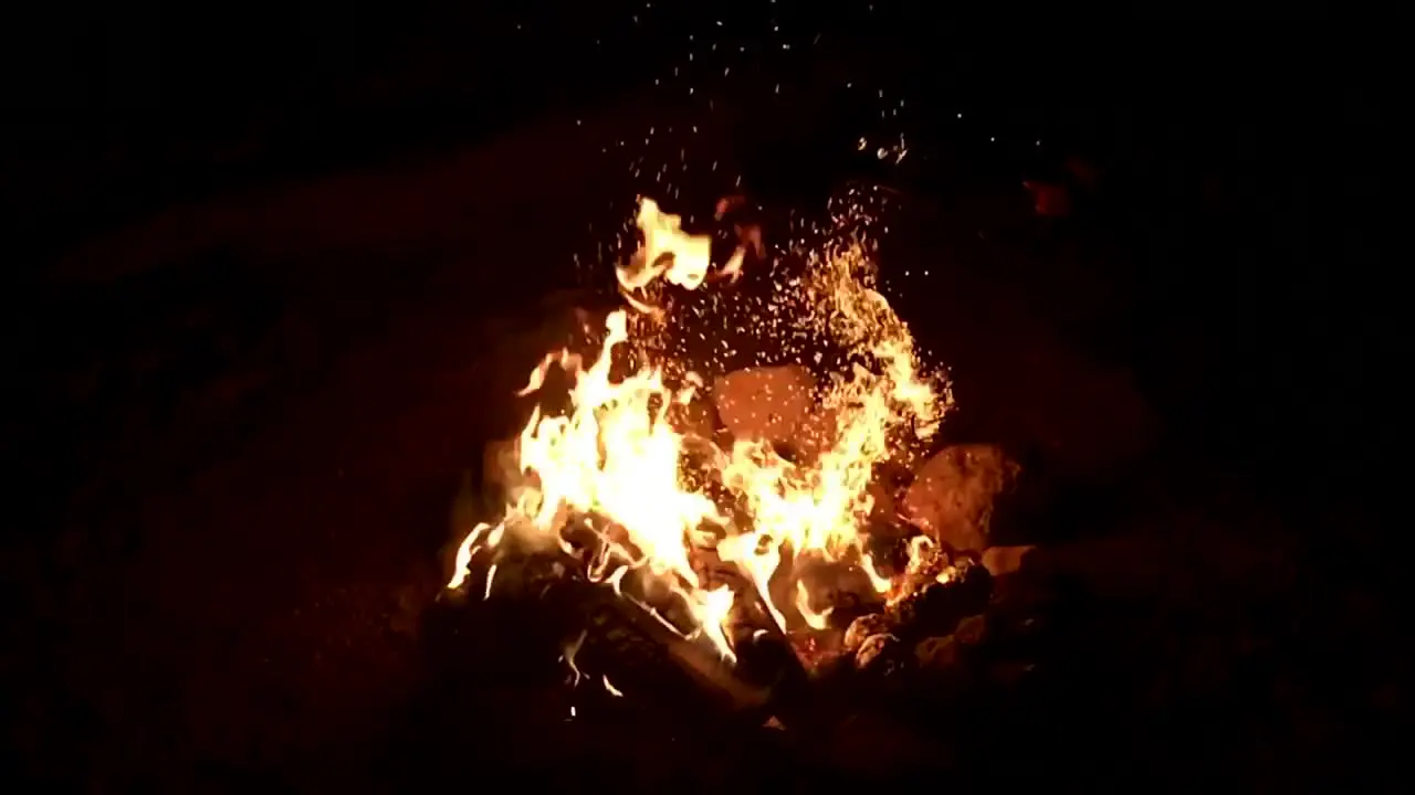 Hand hit campfire burning firewood with shovel causing embers particles to fly at dark night