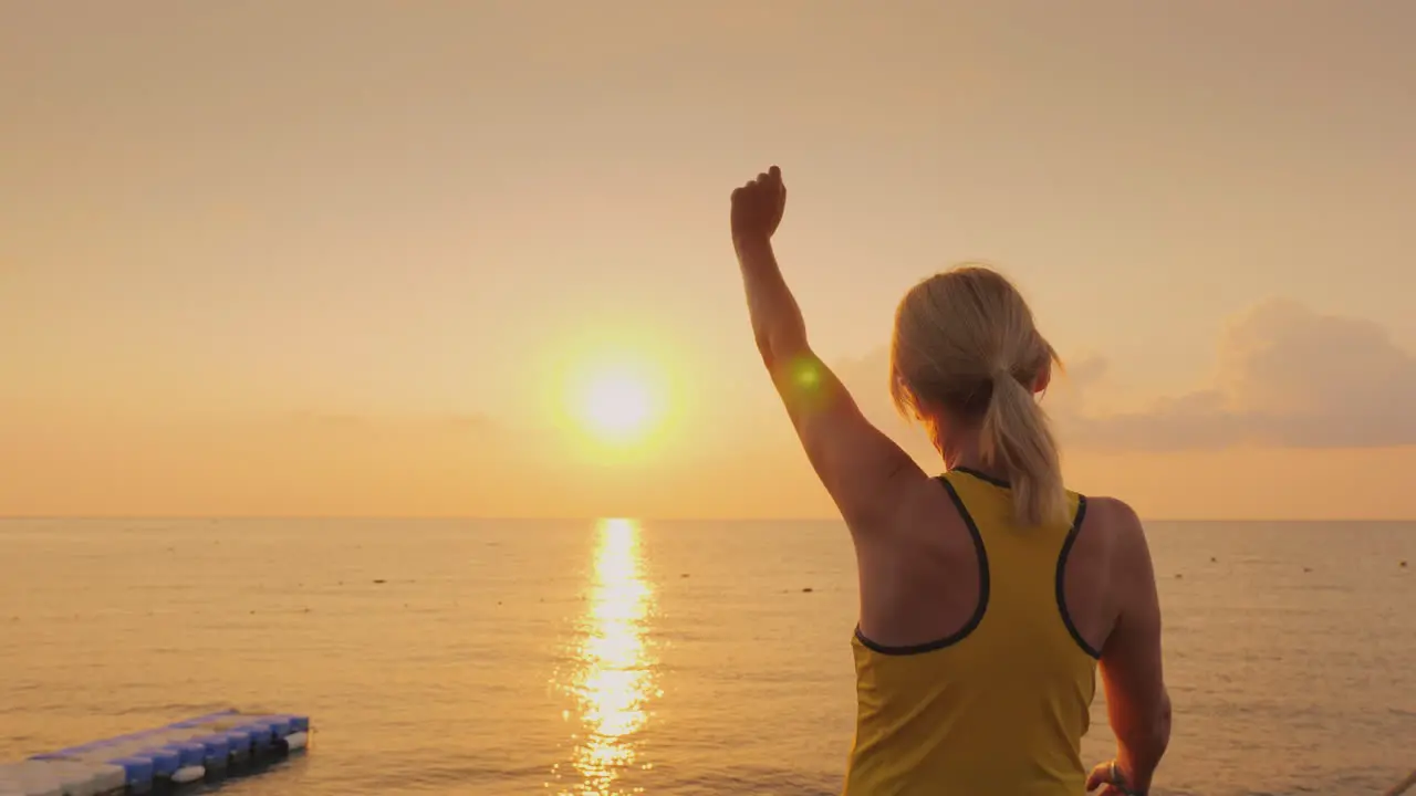 Middle-Aged Woman Warming Up On The Pier On The Background Of A Beautiful Sunrise By The Sea