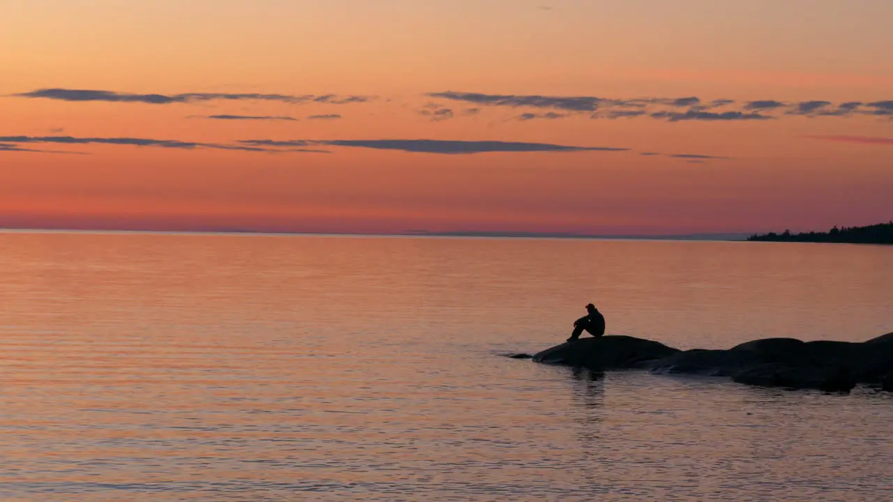 Silhouette of a man on rocky shoreline sitting and enjoying the bright orange sunset