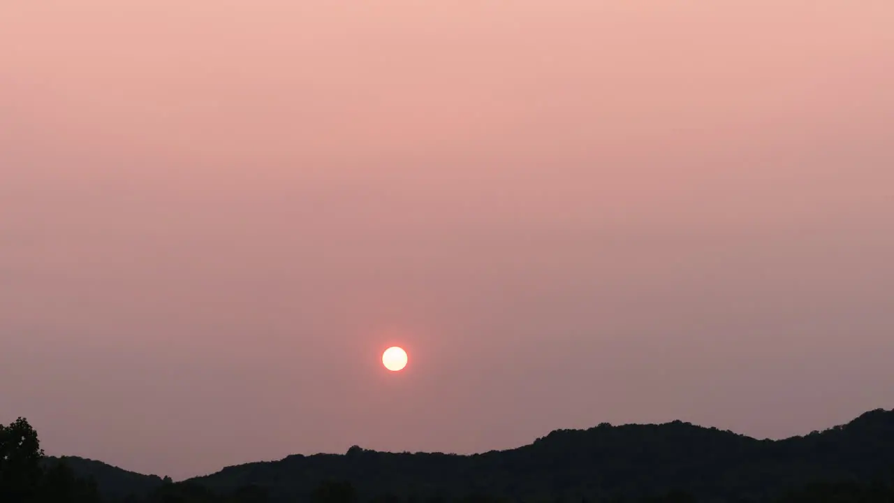 Cloudless hazy sunset time lapse as sun goes behind distant forested hills 4K