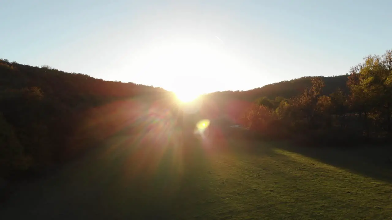 Aerial epic drone shot of sun rays in grass field surrounded by forest at sunset