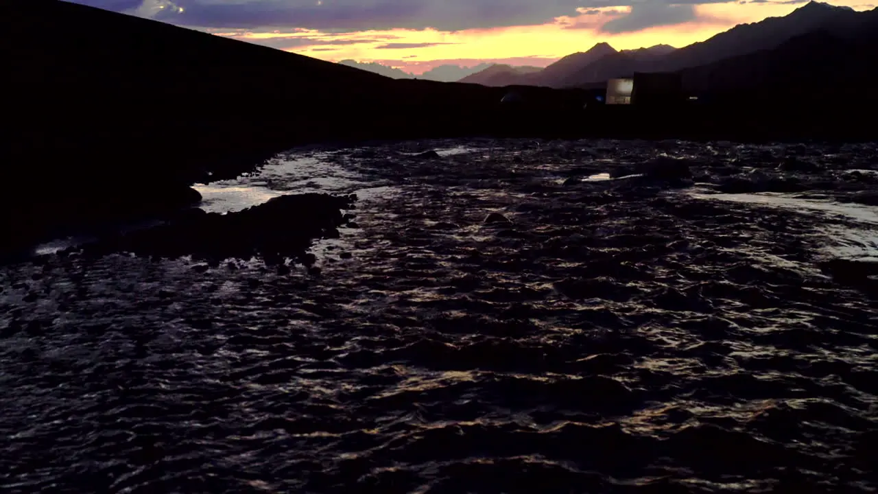 Tilt-up shot from over a river to a mountain scenery with silhouettes of tents base camp in blue and orange sunset