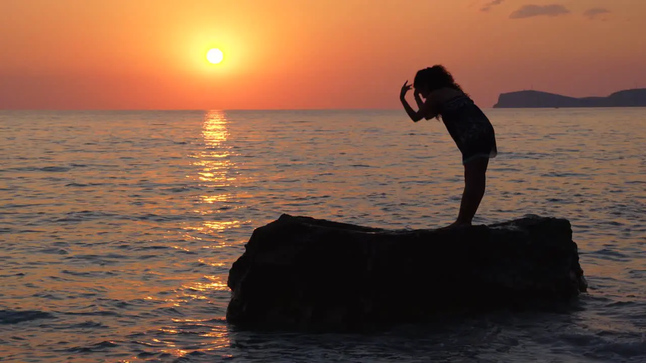 Young woman performing Yoga concentration exercises standing on cliff washed by sea water at beautiful sunset