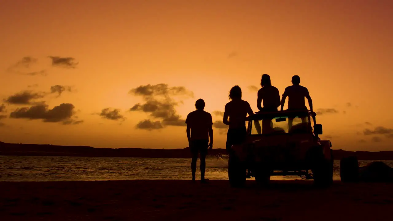 Silhouette showing group of people with quad watching sunset and jumping kiteboarder in ocean static wide shot