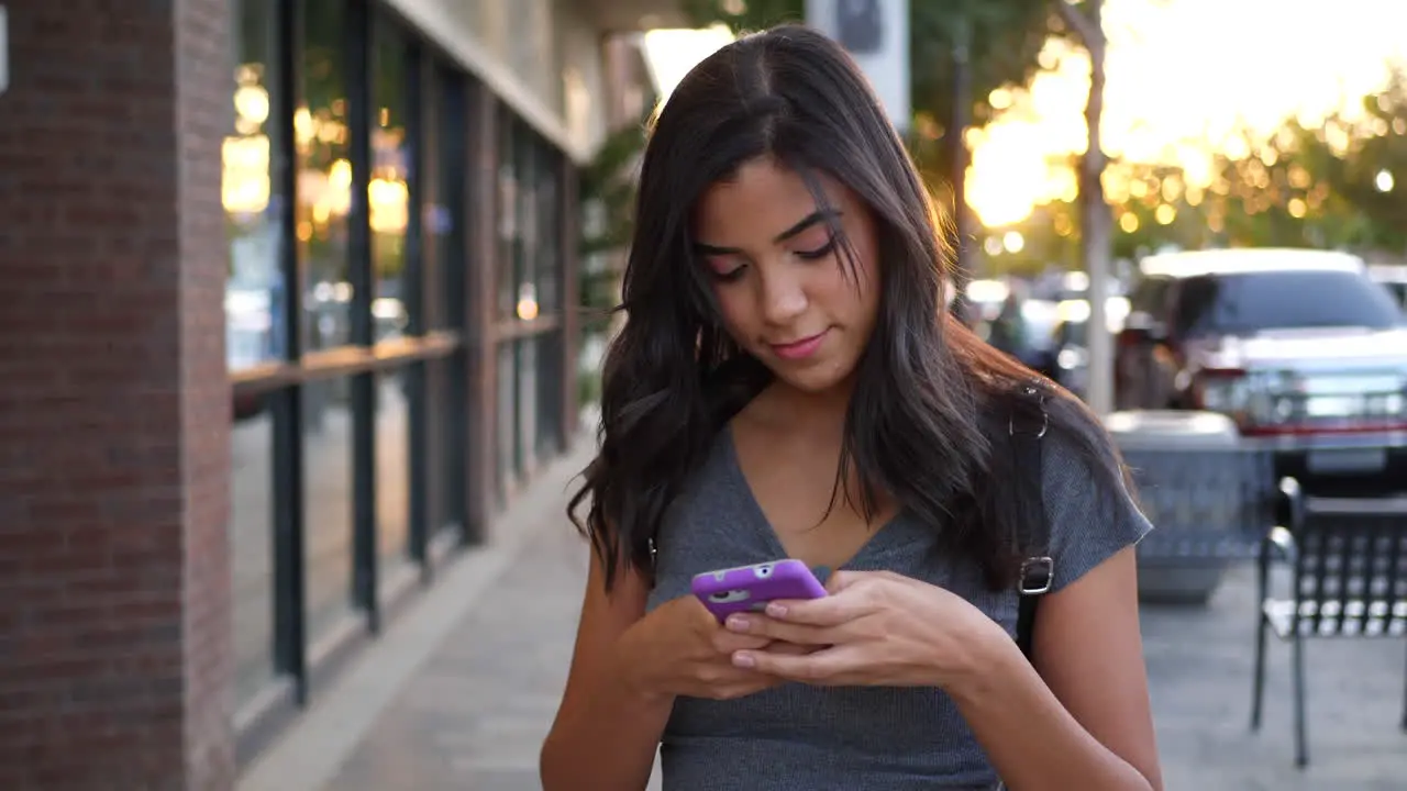 An attractive young hispanic woman walking on urban city streets texting and looking happy at a message on her cell phone SLOW MOTION