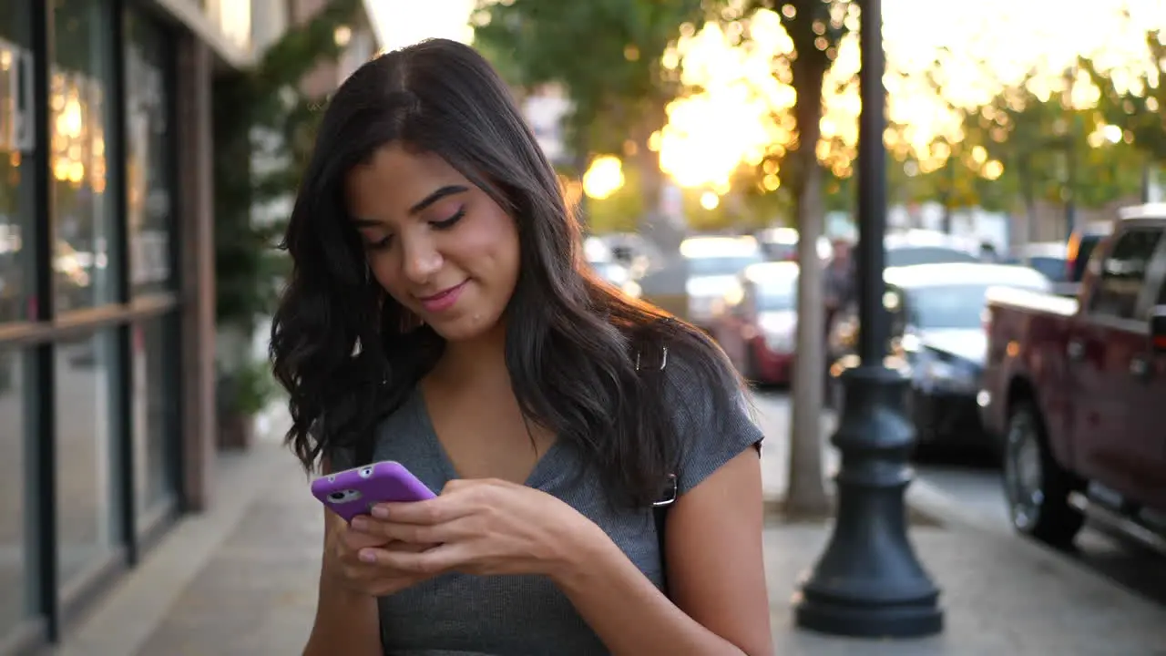 A hispanic woman walking on urban city streets texting and looking happy at a message on her cell phone SLOW MOTION