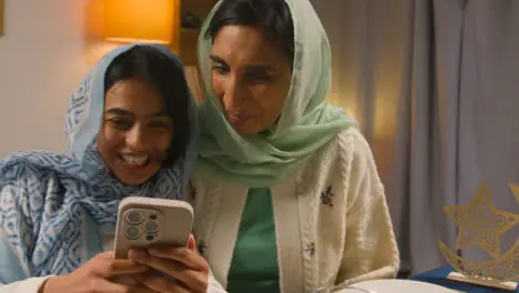 Mother And Daughter Laughing At Mobile Phone Sitting Around Muslim Family Table At Home Eating Iftar Meal Breaking Daily Fast During Ramadan