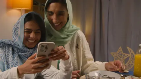 Mother And Daughter Laughing At Mobile Phone Sitting Around Muslim Family Table At Home Eating Iftar Meal Breaking Daily Fast During Ramadan 1