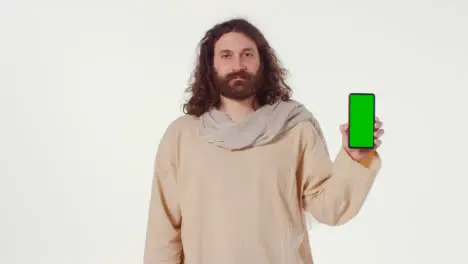 Portrait Of Man Wearing Robes With Long Hair And Beard Representing Figure Of Jesus Christ Holding Green Screen Mobile Phone