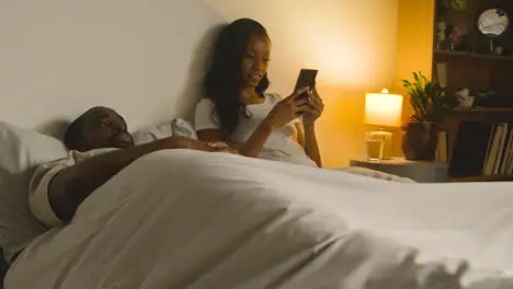 Couple At Home At Night Lying In Bed With Woman Looking At Mobile Phone