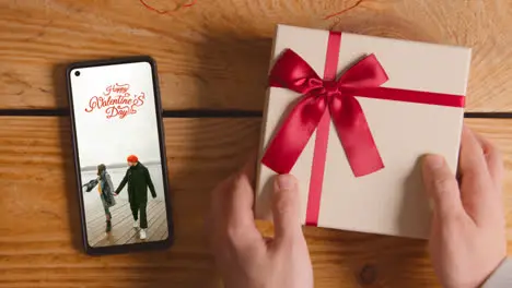 Close Up Of Hand Putting Valentine's Day Gift Next To Mobile Phone With Valentines Message On Screen