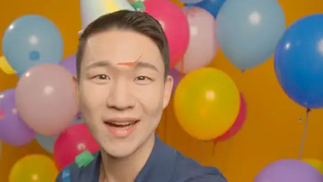 POV Studio Portrait Of Man Taking Selfie Wearing Party Hat Celebrating Birthday Surrounded By Balloons