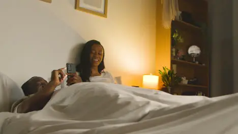 Young Couple At Home At Night Lying In Bed Looking At Mobile Phones Together 1