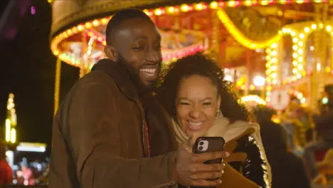 Couple Standing By Fairground Roundabout On London South Bank Making Video Call On Mobile Phone 