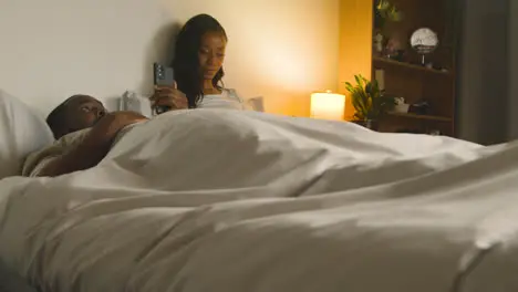 Young Couple At Home At Night Lying In Bed Looking At Mobile Phones Together