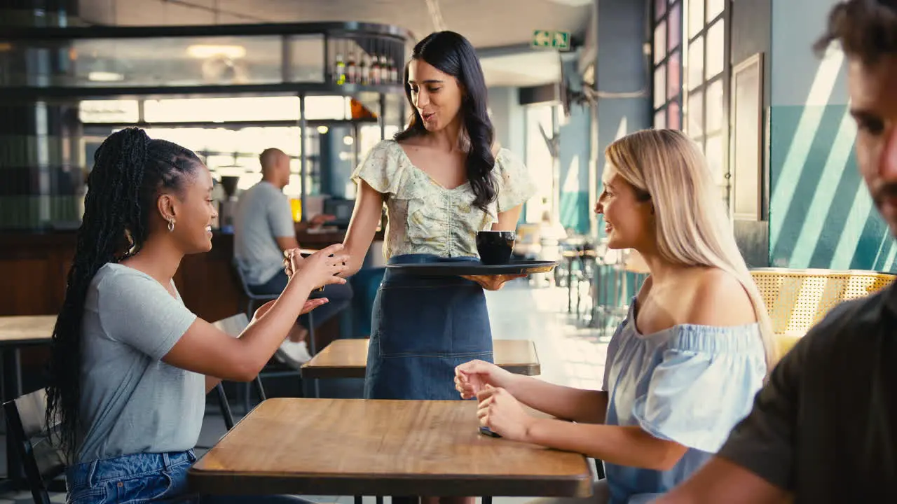 Woman In Coffee Shop Meeting Friend Paying Bill With Contactless Mobile Phone Payment