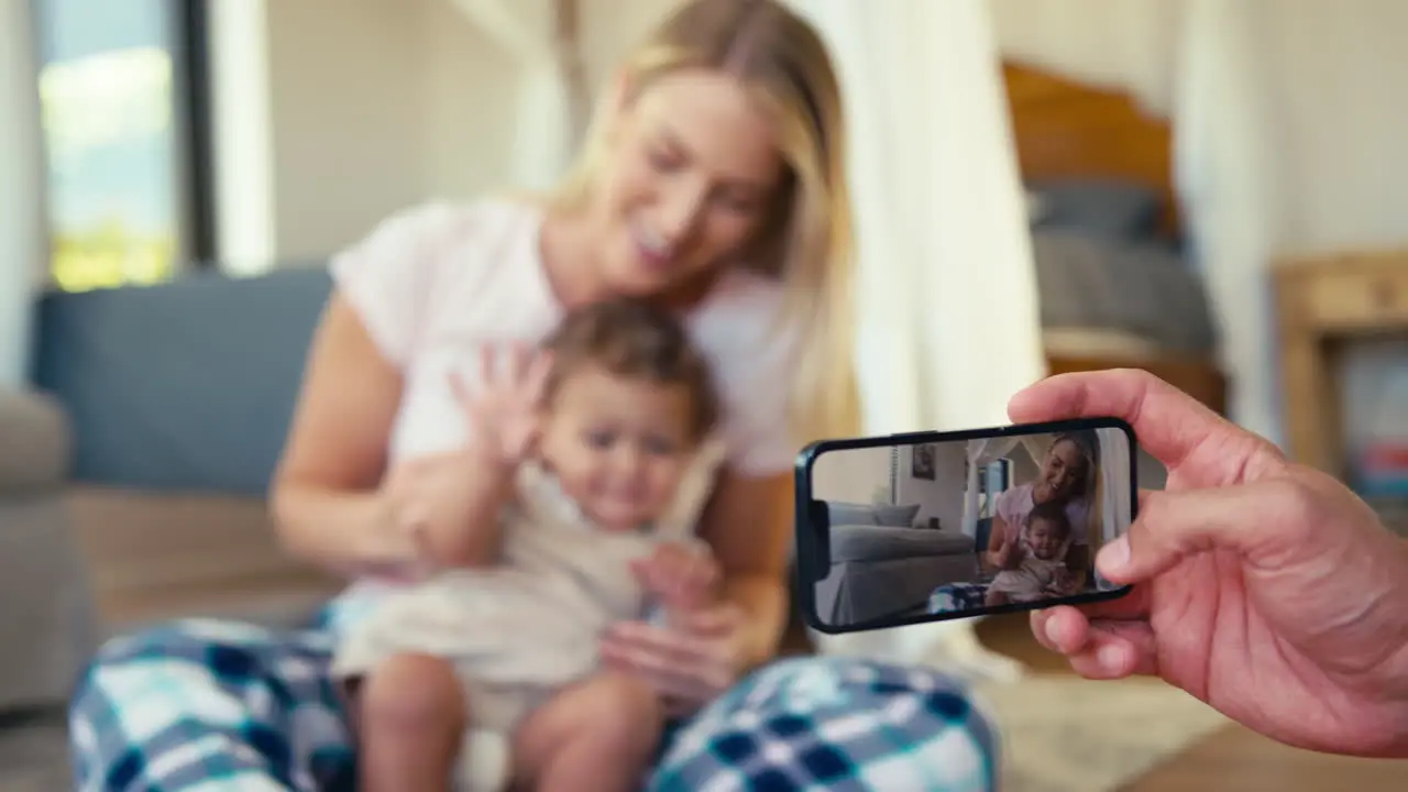 Father Recording Video Of Mother And Son Sitting On Sofa At Home On Mobile Phone