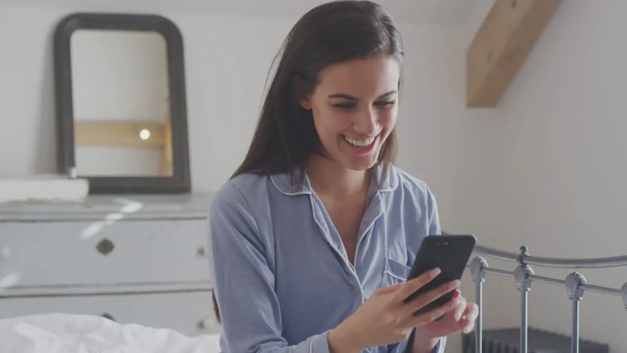 Woman With Mobile Phone Wearing Pyjamas Having Video Chat Sitting On Bed