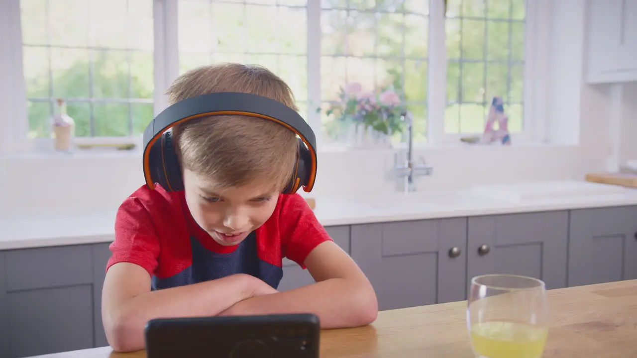 Boy At Home In Kitchen Watching Movie Or Show On Mobile Phone Wearing Wireless Headphones