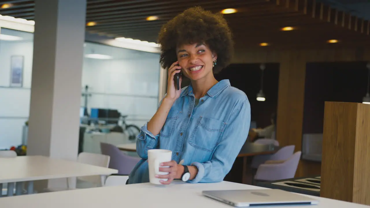 Smiling Businesswoman Standing In Modern Open Plan Office Talking On Mobile Phone