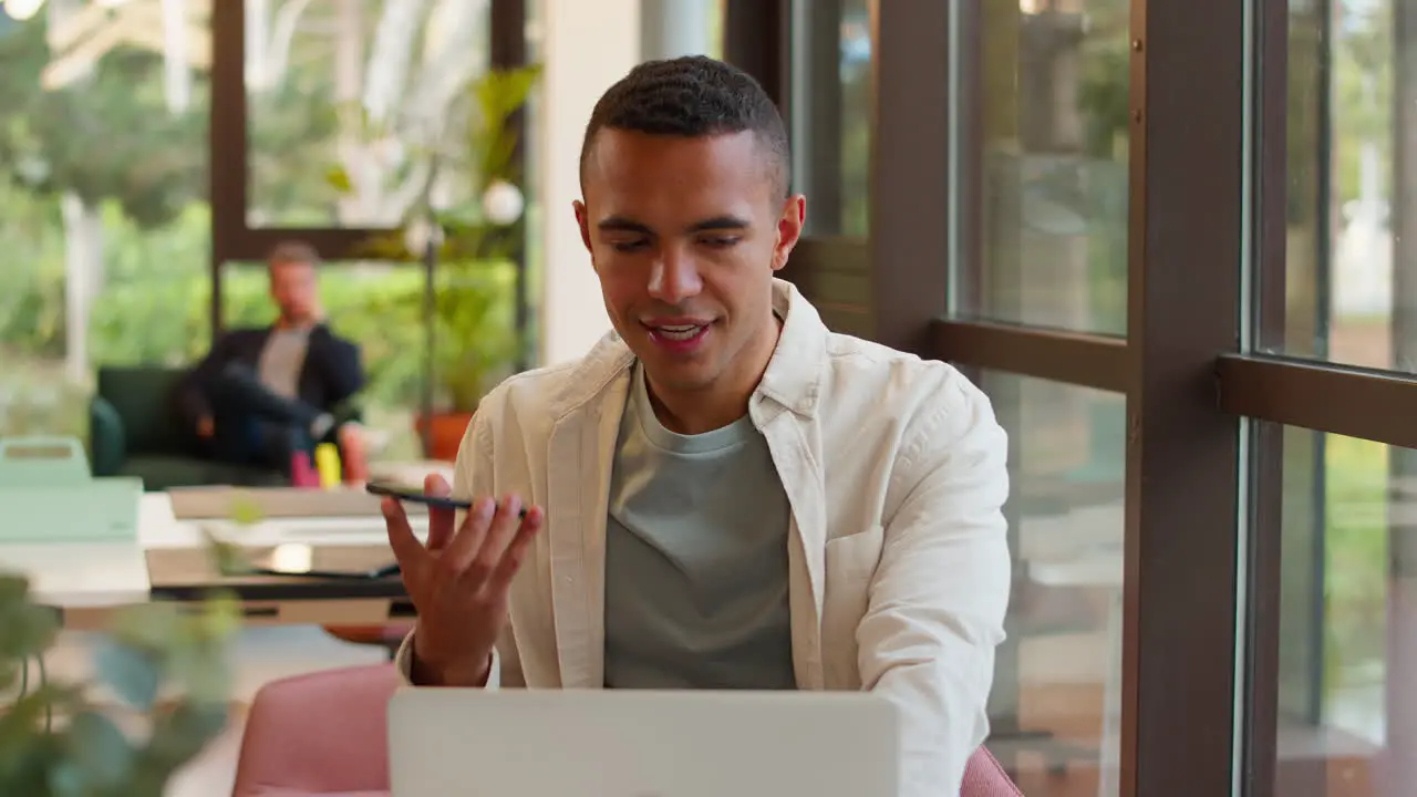 Young Businessman Working On Laptop At Desk In Office Talking Into Mic Of Mobile Phone