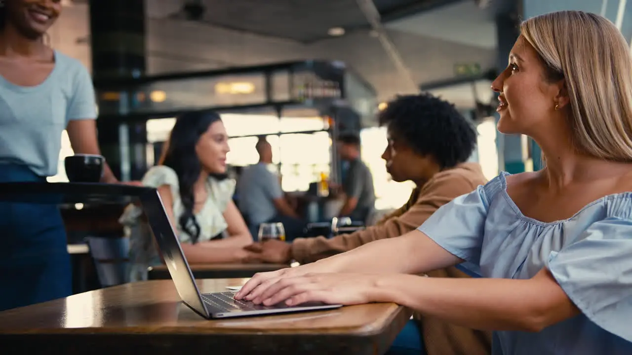 Woman Using Laptop In Coffee Shop Paying Bill With Contactless Mobile Phone Payment