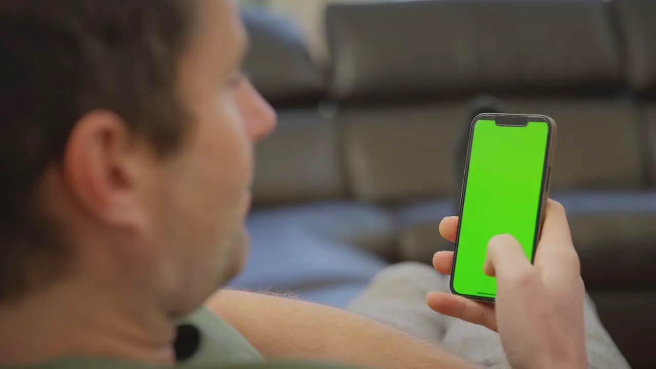 Close up of man wearing pyjamas lying on sofa at home looking at green screen mobile phone shot in slow motion