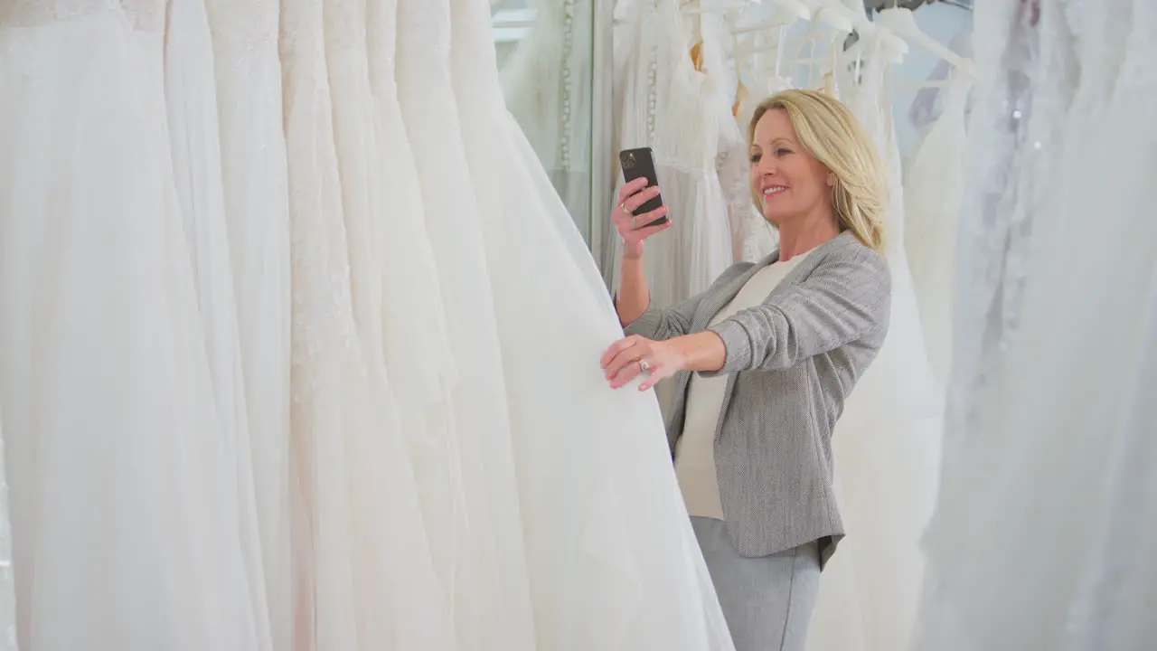 Mature Woman In Bridal Wedding Dress Shop Taking Photo On Mobile Phone