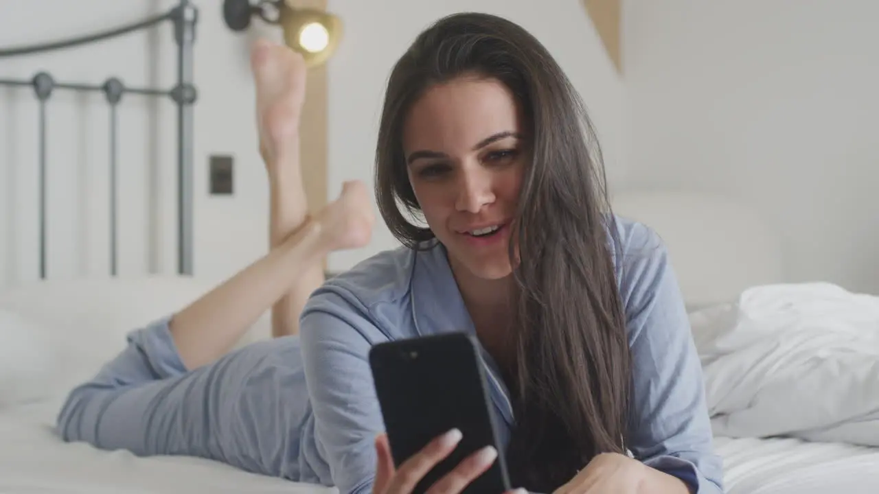 Woman With Mobile Phone Wearing Pyjamas Having Video Chat Lying On Bed